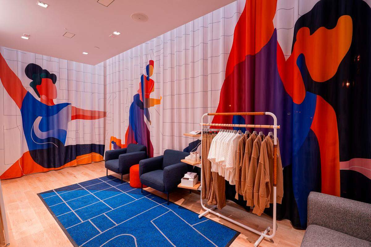 A Nike Live fitting room showcases neon-colored abstract wall art of women stretching in various yoga positions. Blue chairs and a gray couch line the fitting room walls on top of a blue rug. A simple rack of beige sweat suits and white t-shirts hangs in the corner.