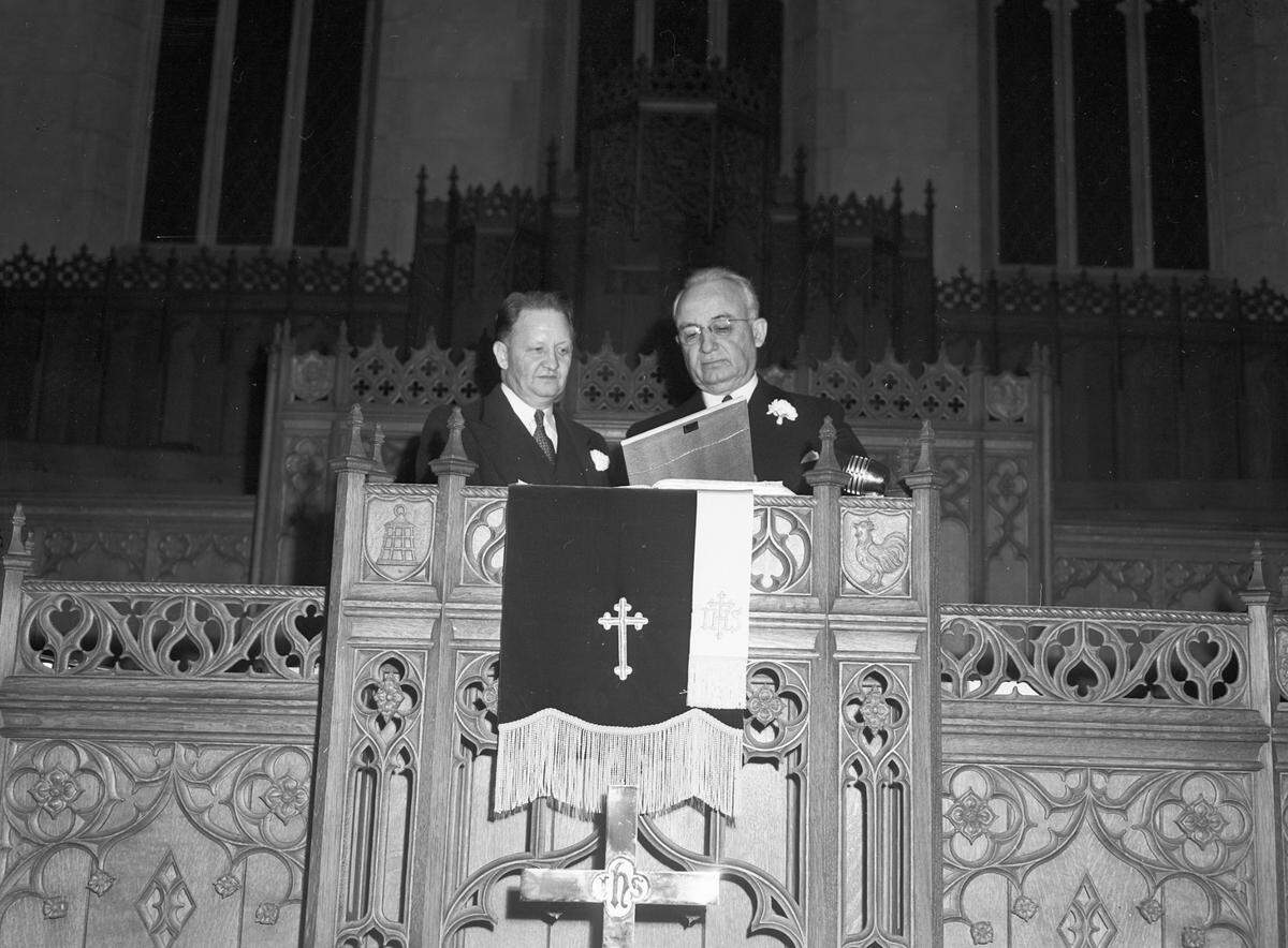 First Methodist Church dedication in 1945. The Rev. Warren Johnston, left, and Bishop A. Frank Smith of Houston, Texas. Johnston is the pastor. He is wearing a suit with a carnation pinned to the lapel. Bishop Smith is presiding bishop of the Central Texas Conference who accepted the building for the denomination. He is dressed in a suit with a carnation pinned to his lapel. The men are at altar examining the framed, canceled note which lifted the last of the congregation's indebtedness.