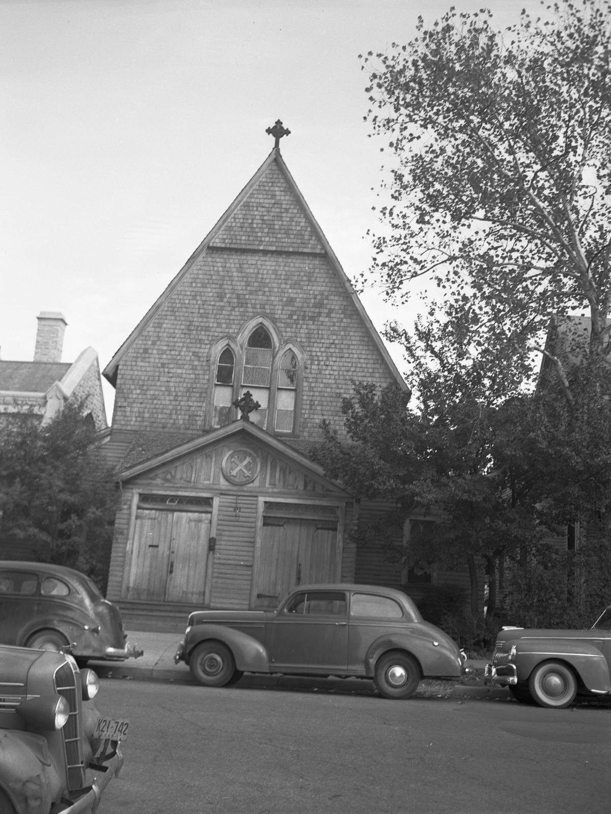 A street view of the little frame parish house of Saint Andrew's Episcopal Church, with several automobiles parked on the street, in front of the church.