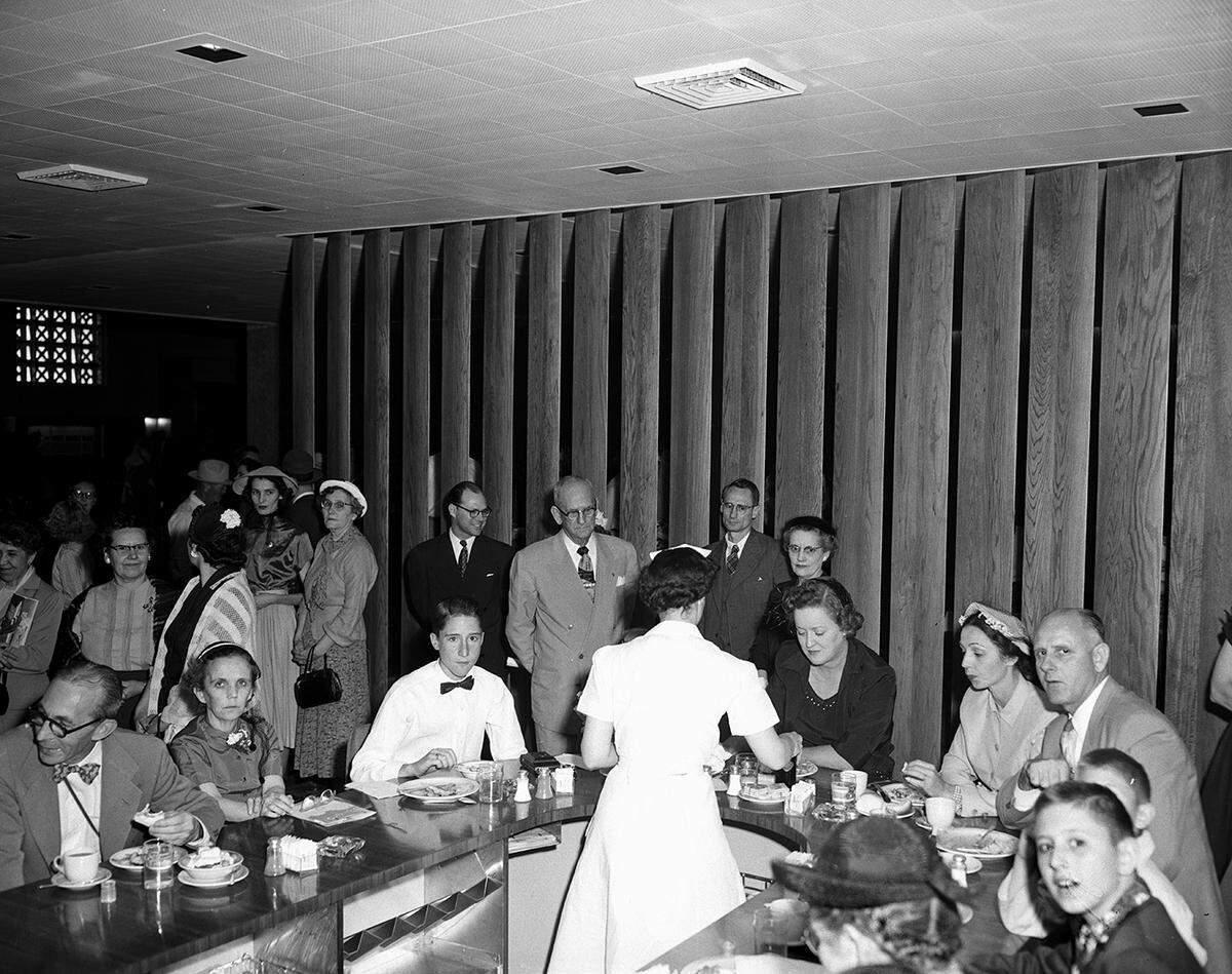 A group of people are seated at a round counter being served food by a waitress. There are people standing in the background.