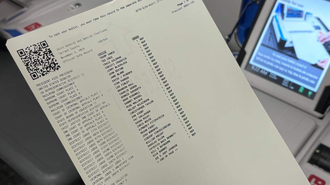 A white piece of paper with lines of text dominates the field. A square QR code sits at the top left corner of the paper. A finger is pointing at the paper from the right side of the frame, and a computer screen can be seen in the background. 