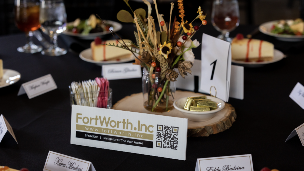 A luncheon table is decorated with a vase of fall-themed flowers, a table number, name cards, plates full of salad, and a "Investigator of the Year" card with Fort Worth.Inc listed as the sponsor.