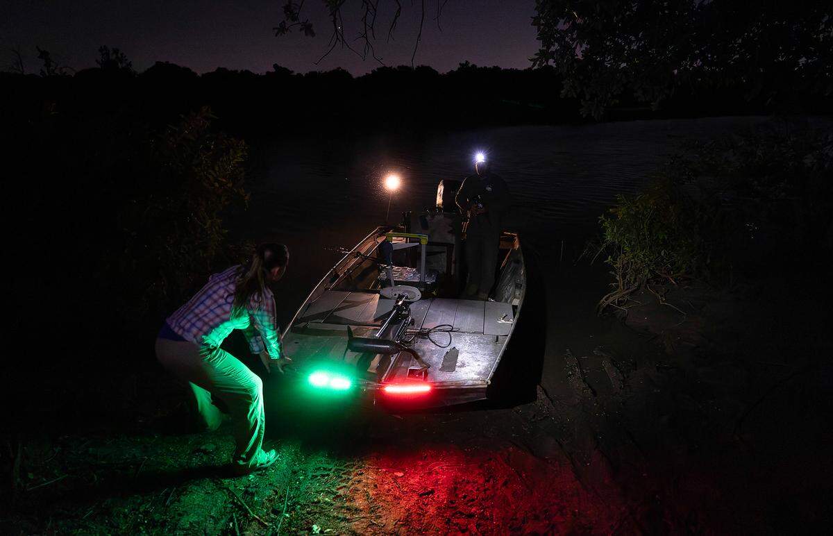 A woman pushes a boat into the water. It is dark except for a few sparse lights.