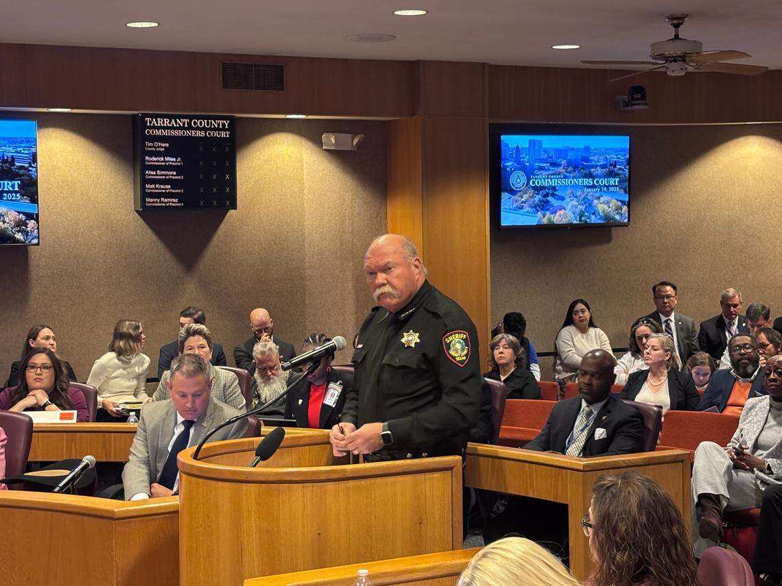 A large white man in a black law enforcement uniform speaks at a dais. The man is bald with a white mustache. People sit in rows of seats behind him.