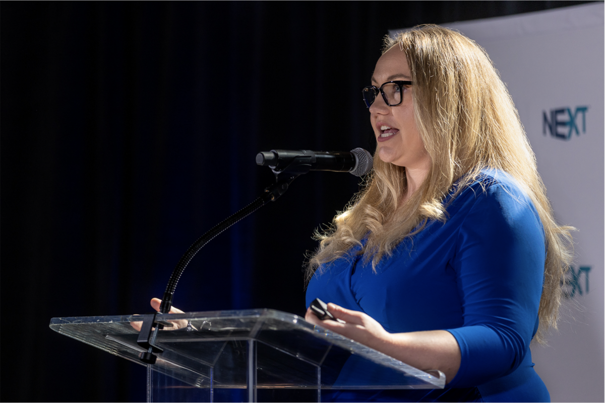 Jennifer McDonald wears a blue business dress and stands at a glass podium with a microphone. Her hands are placed on the podium as she speaks.