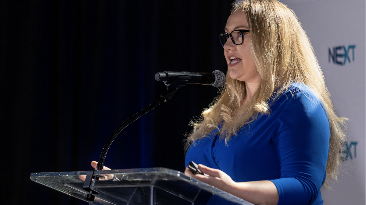 Jennifer McDonald wears a blue business dress and stands at a glass podium with a microphone. Her hands are placed on the podium as she speaks.