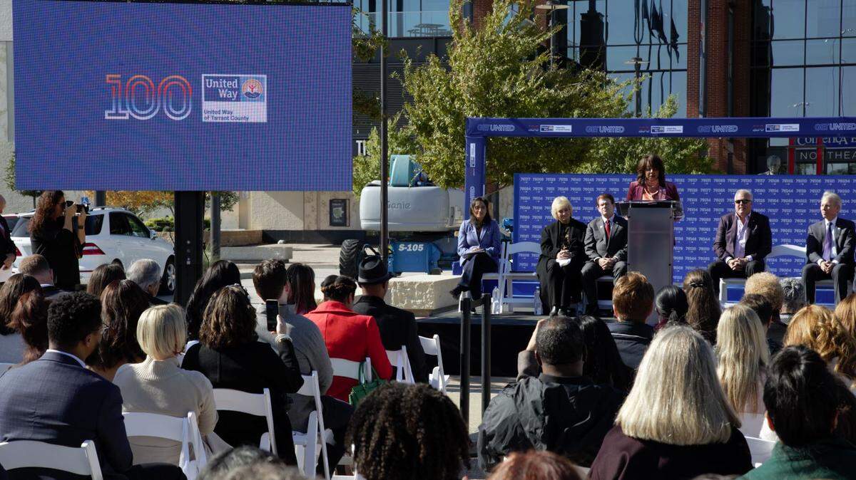 A woman speaking on a stage to a crowd on a sunny day. 