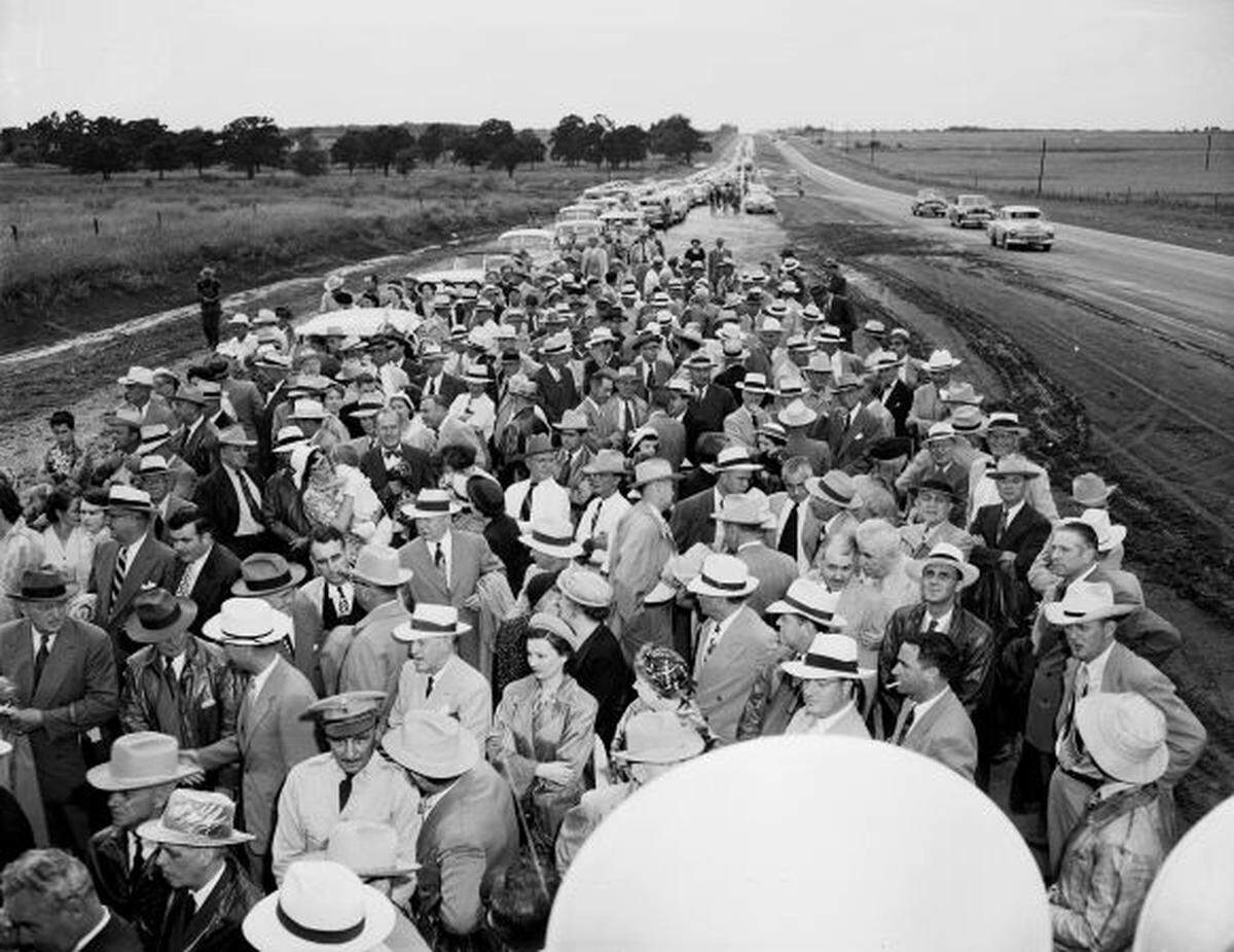 A photo shot from above of a large group standing along the highway, with cars parked in the background.