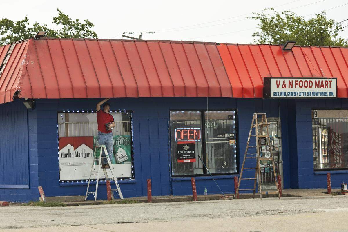 A man on a latter reaches above him to paint a red tin roof of a corner store.