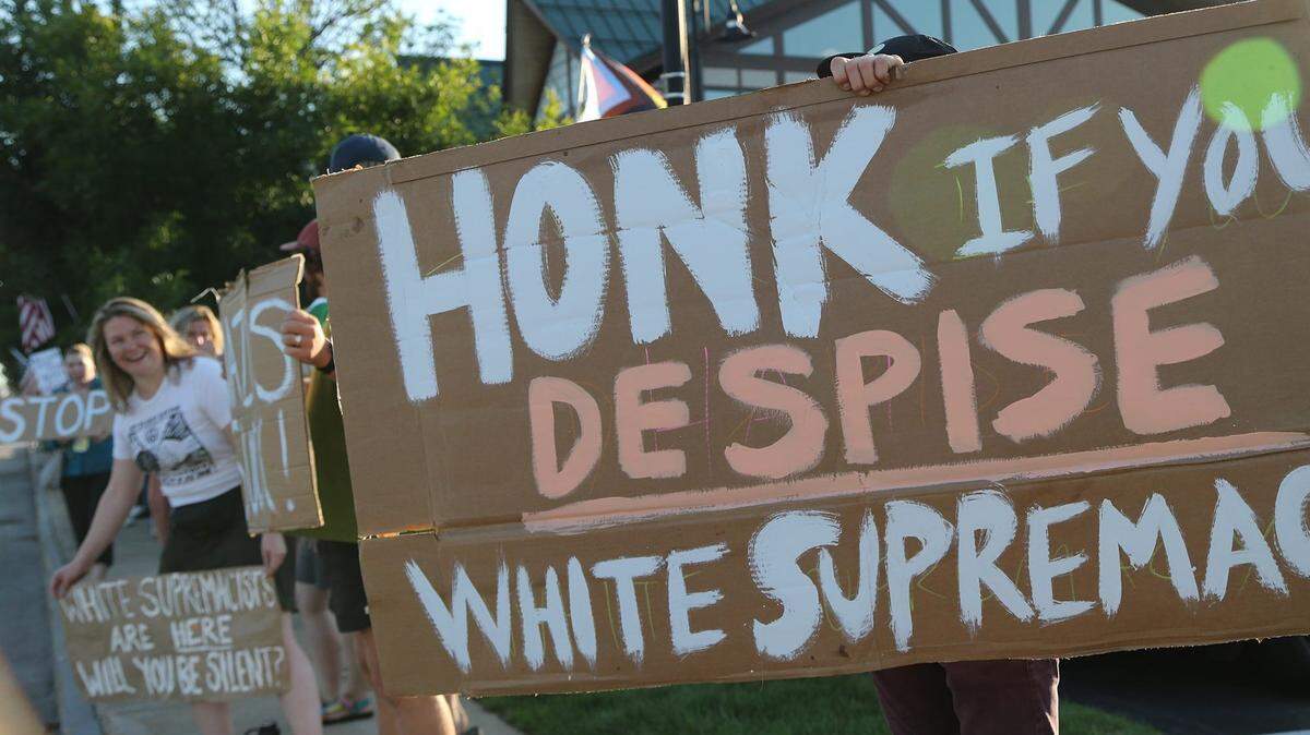 People standing along a street hold signs that say, Honk if you despise white supremacy. 