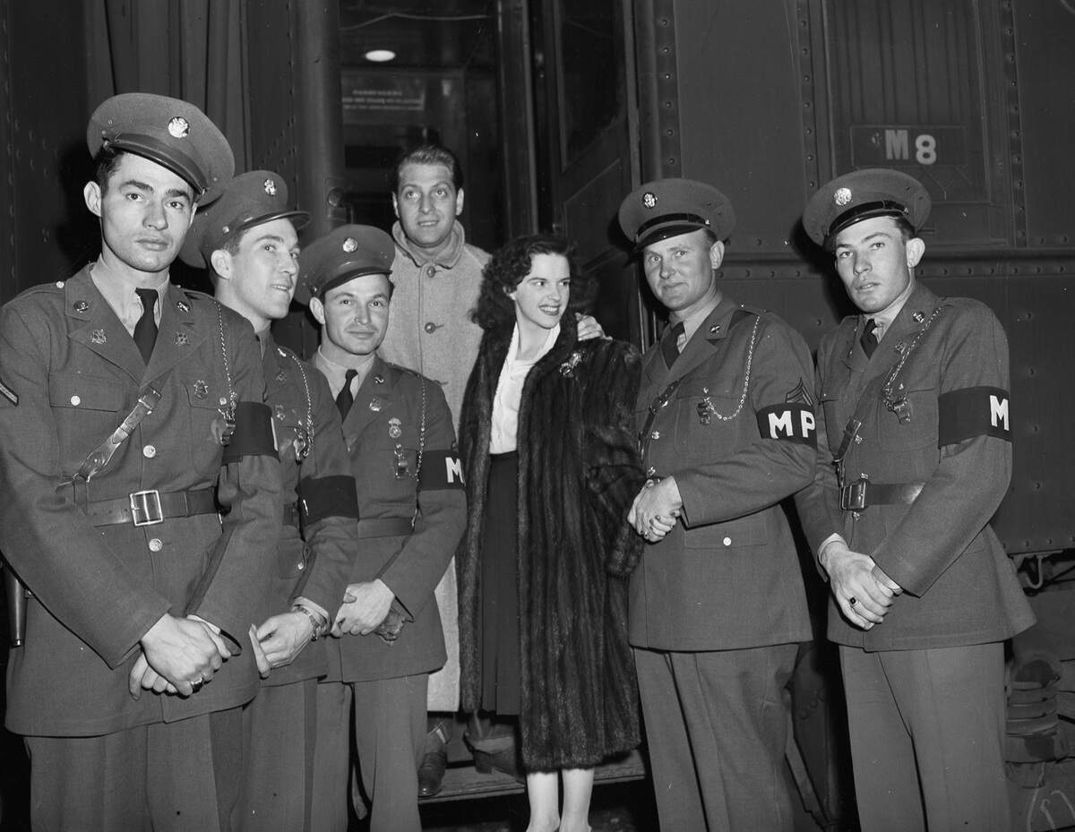 The officers are dressed in their military uniforms. Mr. Rose, a composer and director, is wearing a buttoned winter coat. Miss Garland, famous for her role in the Wizard of Oz, is wearing a button-up blouse, skirt, a fur coat, and a brooch. 