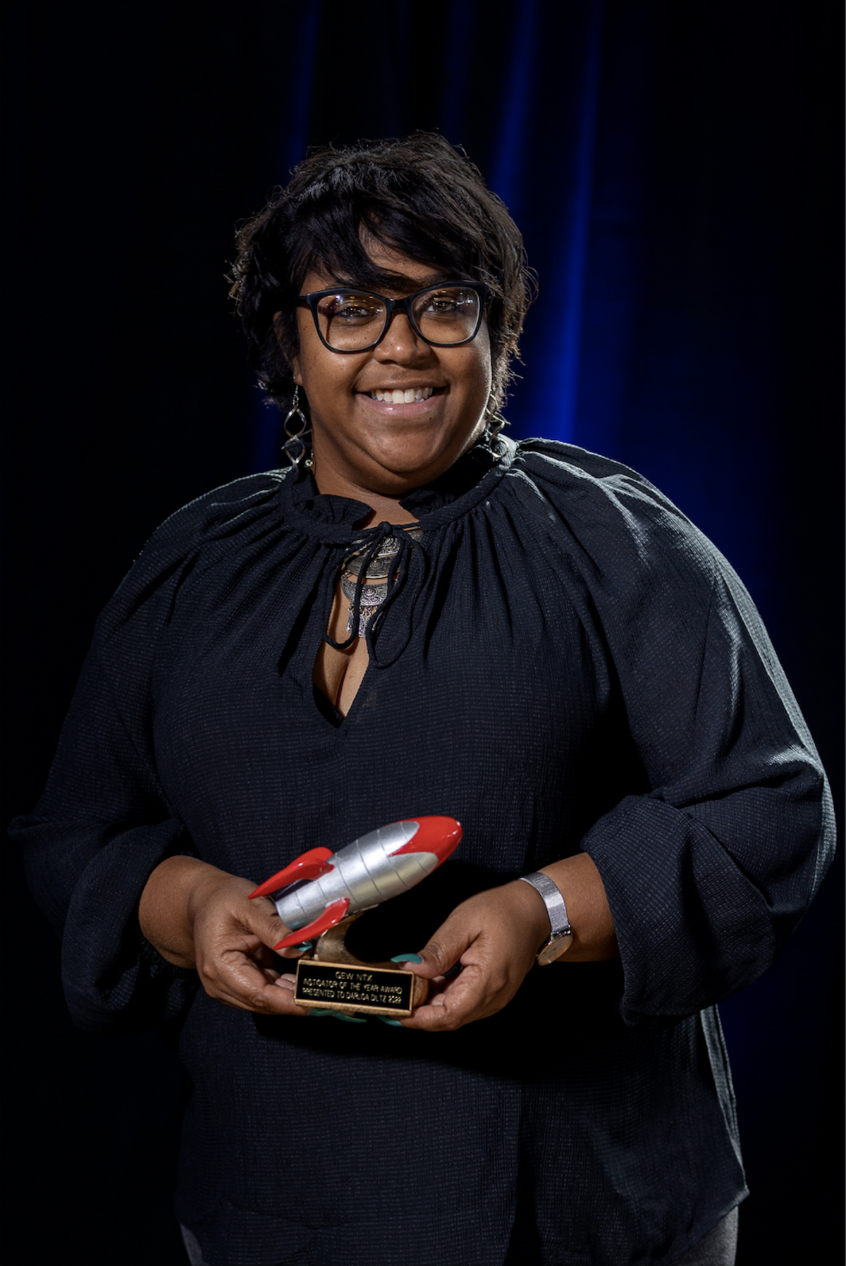 Darlisa Diltz smiles and poses holding a trophy with a red and silver rocketship on it to celebrate winning Instigator of the Year.