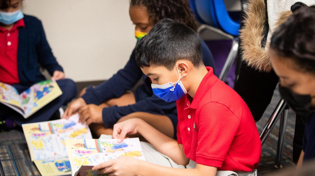 A boy in a red polo shirt and a face mask sits on a floor reading a book.