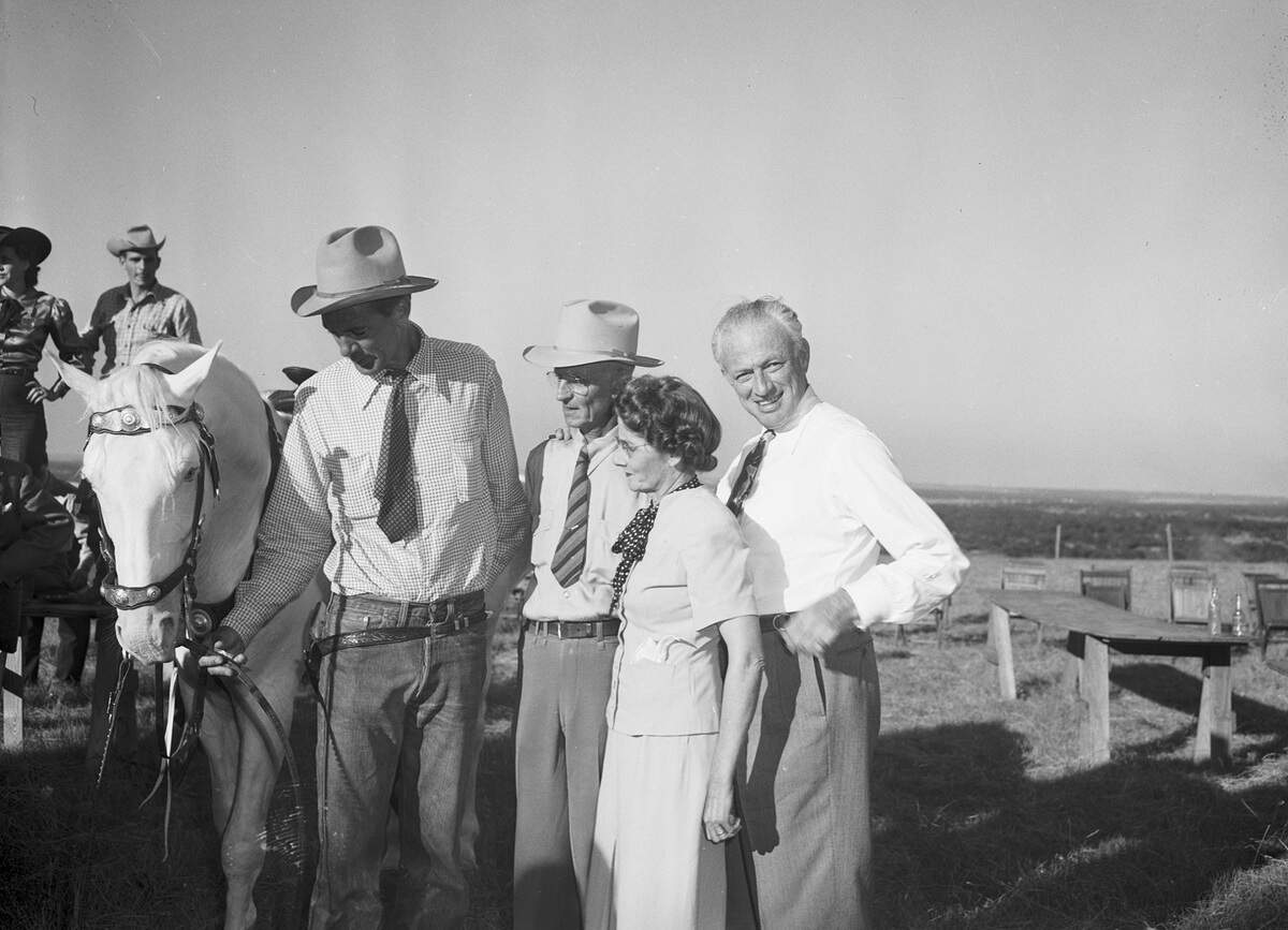 Beside Cooper, at the far left, is a horse. Cooper is wearing a long-sleeve collared shirt, a necktie, a holster, a cowboy hat and jeans. Mr. Wilkinson is wearing a long-sleeve collared shirt, striped necktie, cowboy hat, trousers and eyeglasses. Mrs. Wilkinson is wearing a short-sleeve blouse, a scarf, a skirt and eyeglasses. O'Donnell is wearing a long-sleeve collared shirt, necktie and trousers.