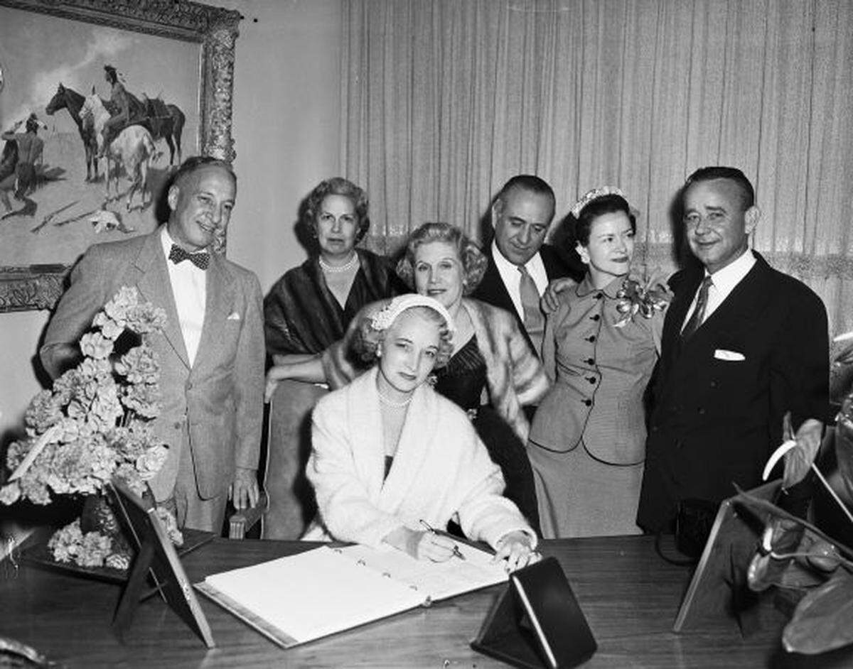 Mrs. Carter is seated at a desk appearing to sign a guest book while others stand around her in a fancy lounge.