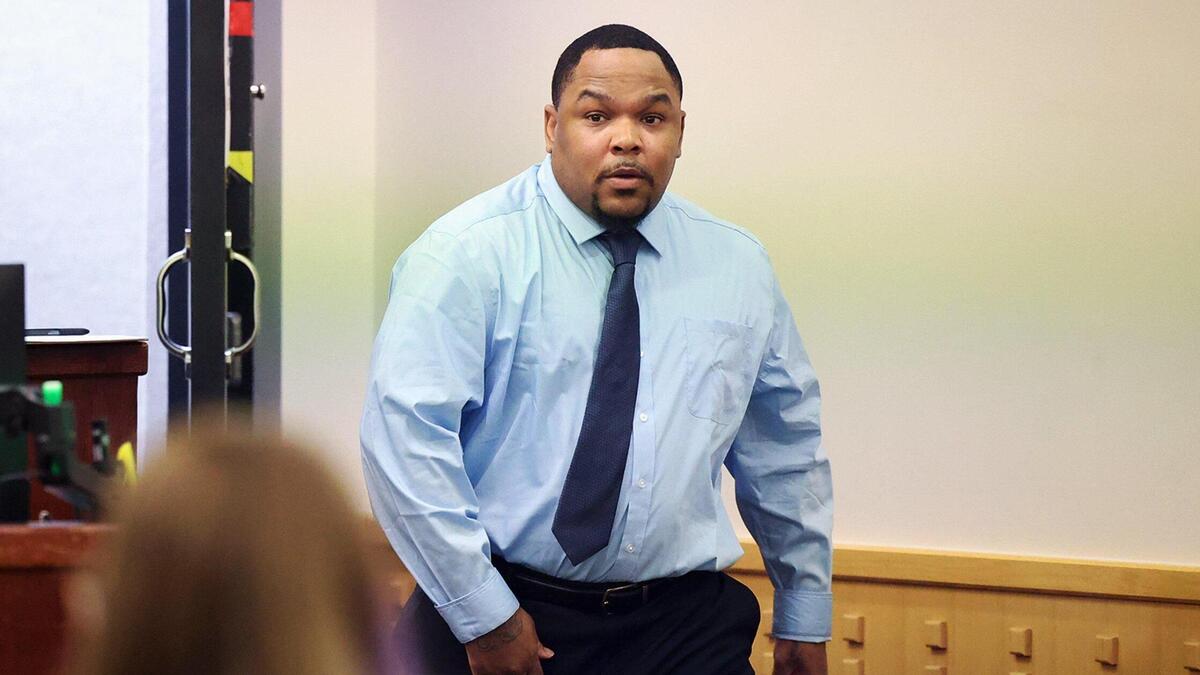 A man wearing a light blue shirt and dark blue tie walks into a courtroom, a slightly open door behind him.
