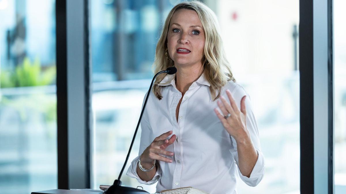 A blonde lady in a white blouse speaking behind a podium.