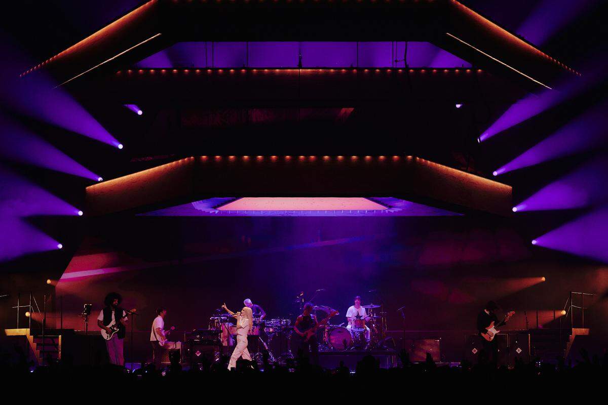 A wide shot of a stage, lit from above, with three polygons hanging above the stage. Seven musicians play onstage.