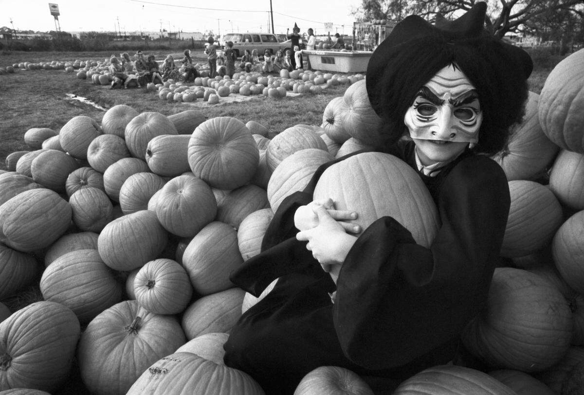 A child is seen at a pumpkin patch, dressed in a witch costume with a creepy mask, with her arms wrapped around a pumpkin.