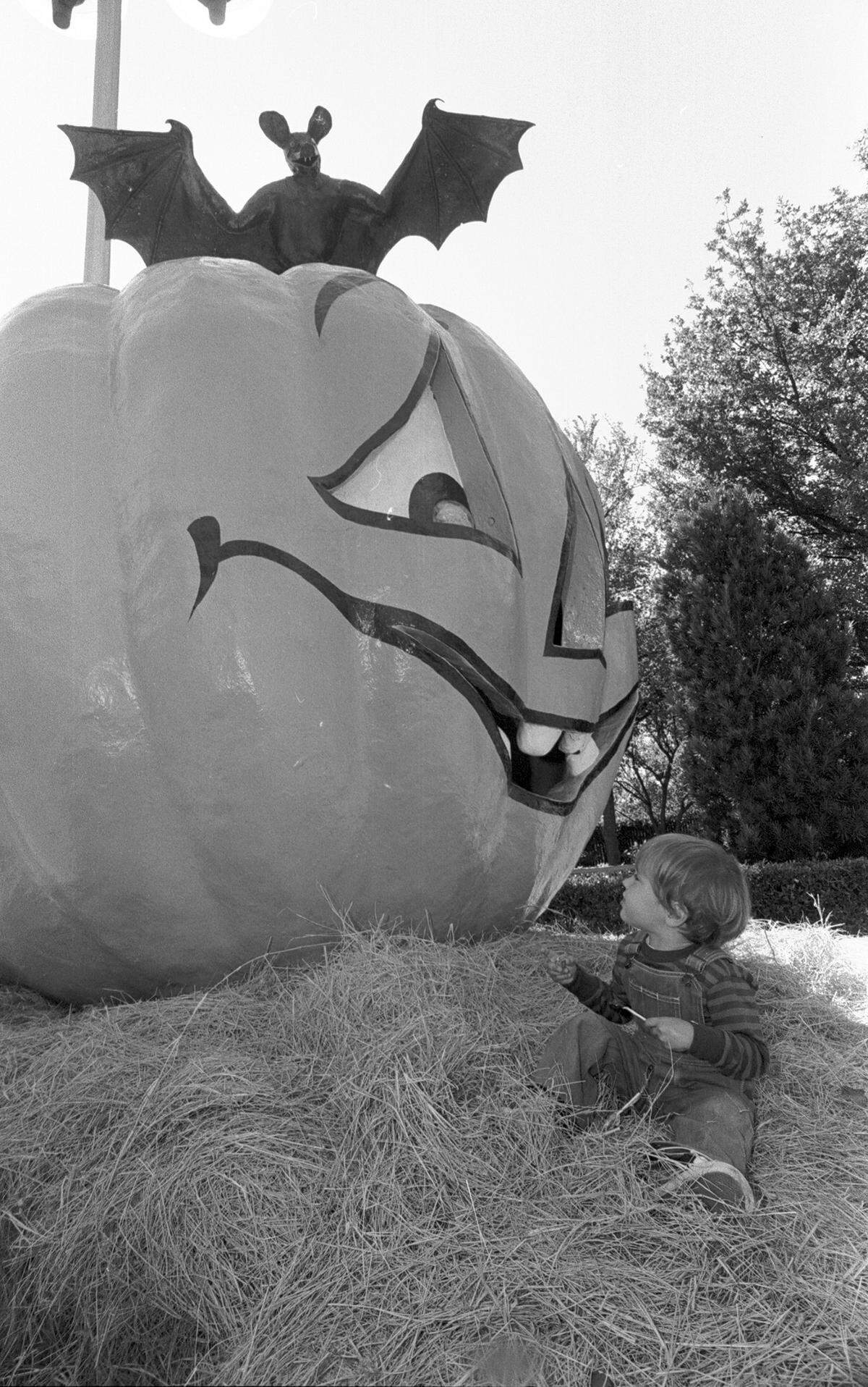 A toddler sits in hay, looking up at an oversized fake pumpkin with a smiling face on it. 