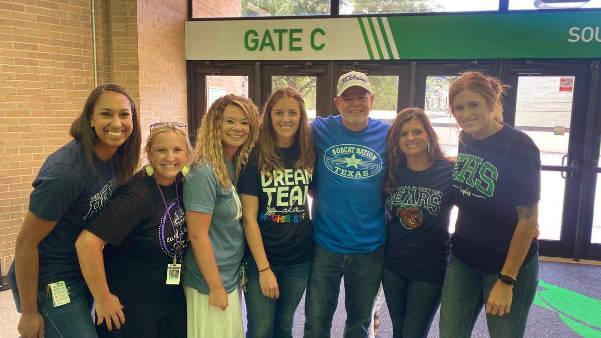 Six women stand with their former high school basketball coach in concourse of auditorium.
