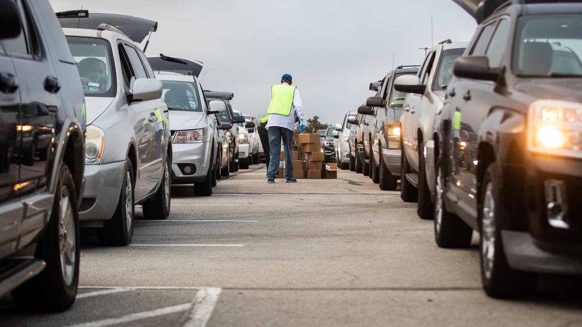 Two men stand with cardboard boxes in between two lines of vehicles, all with their trunks open, in a parking lot.