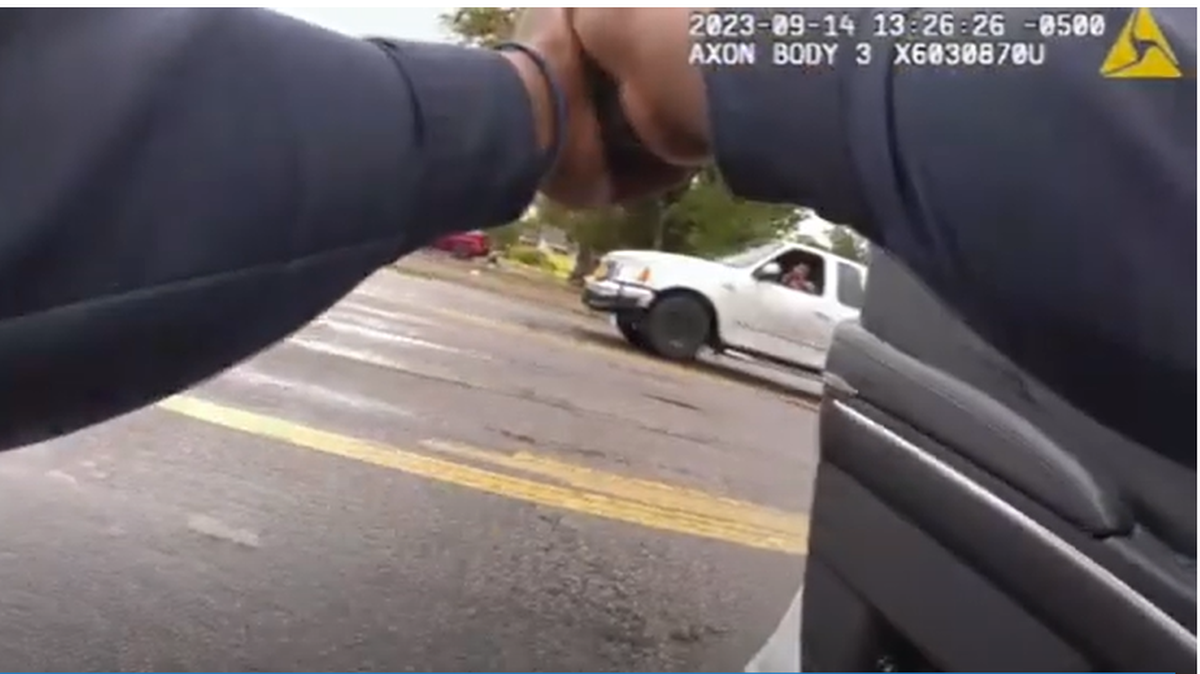 A body camera screenshot showing a white truck blurring by as a police officer raises his gun