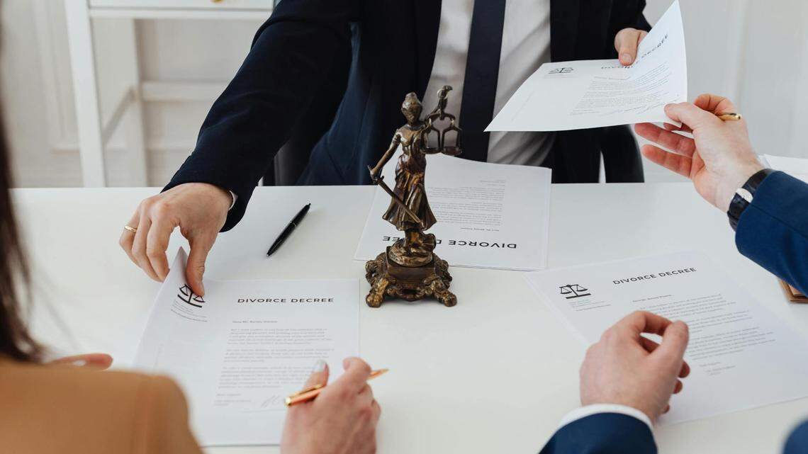 A stock photo of a man and a woman's hands are seen holding divorce papers to give to a lawyer. a paperweight of Lady Justice sits on the desk table.