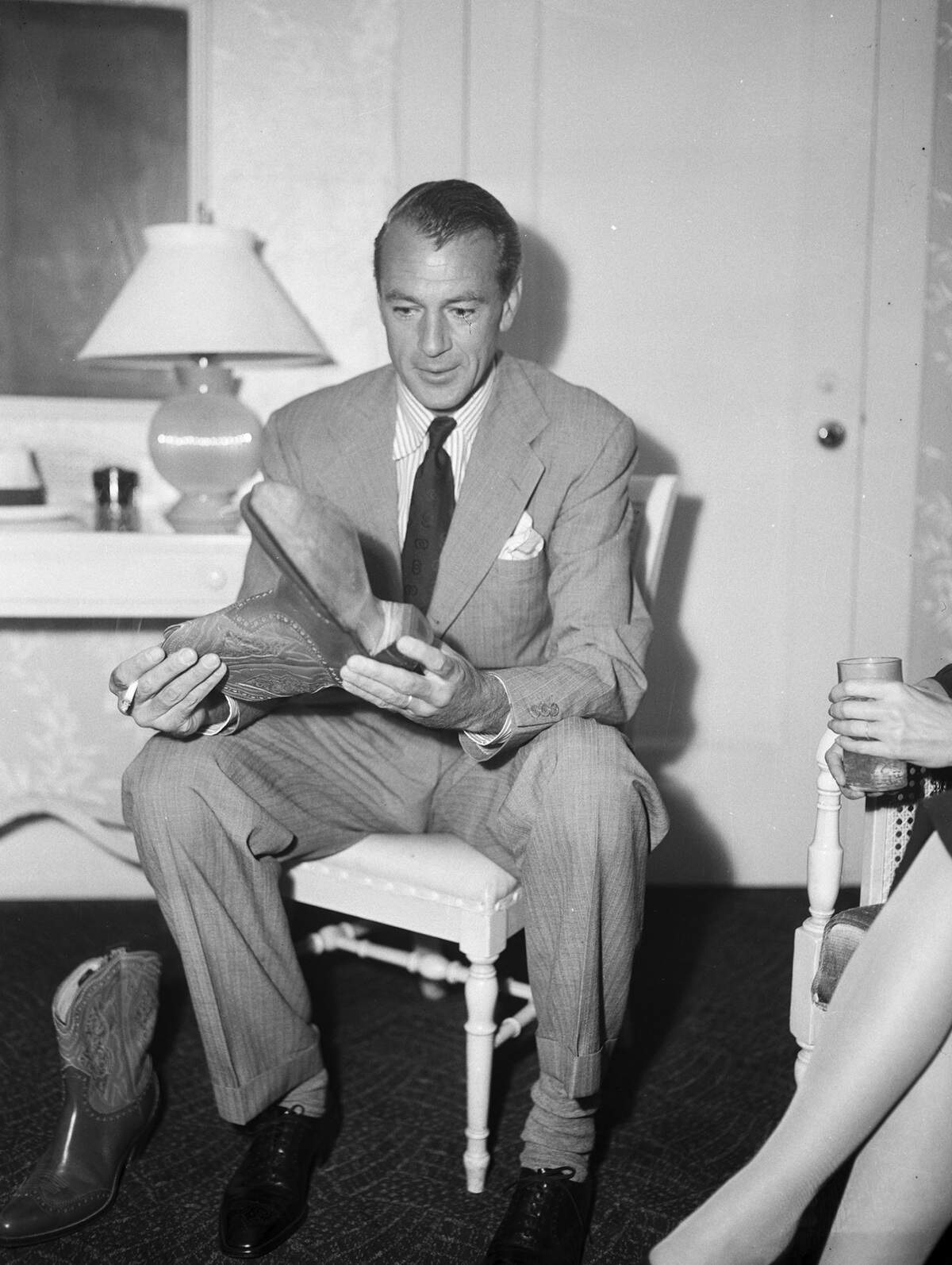 An unidentified woman is sitting at the far right (partially obscured). Cooper is wearing a suit and holding one of the boots. The other boot is on the floor beside him, at left.