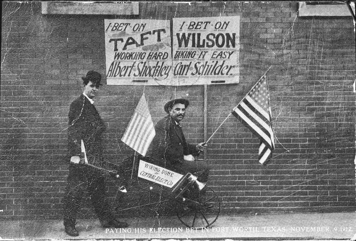 Nov. 9, 1912: Albert Shockley pushing Carl Schilder down Main Street in a wheelbarrow. There is a sign on the brick wall behind them that reads, “I bet on Taft working hard, Albert Shockley” and “I bet on Wilson taking it easy, Carl Schilder”. The caption under the image reads “Paying his election bet in Fort Worth, Texas.