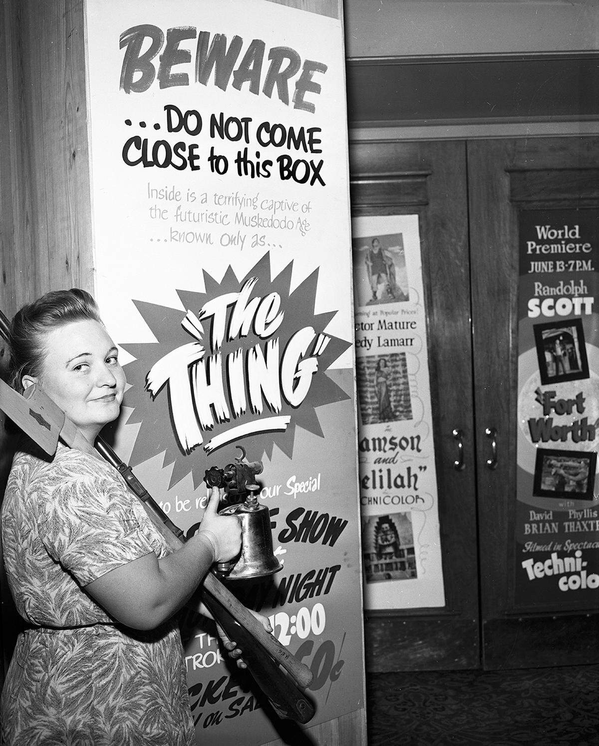 A young woman in a dress poses and smiles in front of a movie poster carrying what appears to be an axe. 