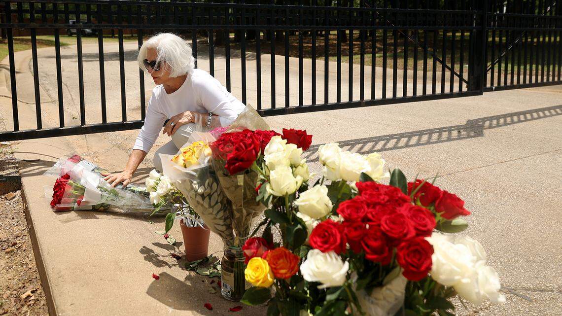 A woman kneels and touches two bundles of roses placed in front of a black iron gate. More roses fill the foreground to the right.