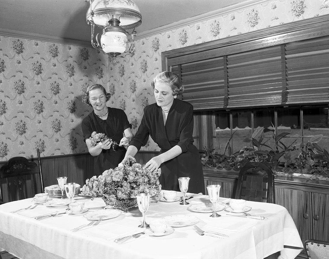 Nov. 1, 1952: Society series on Thanksgiving table settings. Finishing Touches: At the home of Mrs. W.O. Kelly, 1825 Oakland, she is assisted by Mrs. Jack Kelly in putting the last posies in place before the family dines. Damask, imported china and silver set this table.