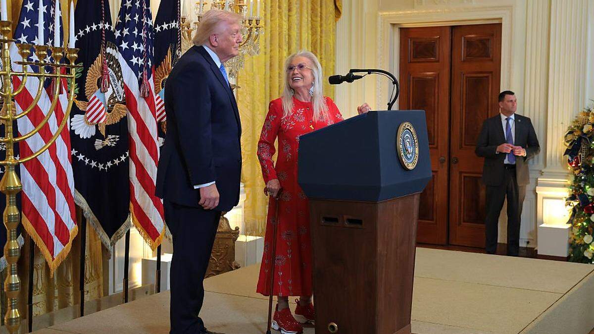 WASHINGTON, DC - DECEMBER 16: U.S. President Donald Trump (L) looks on as Miriam Adelson speaks during a Hanukkah Reception in the East Room of the White House on December 16, 2025 in Washington, DC. Trump hosted attendees to celebrate the holiday and the lighting of the menorah on the third night of Hanukkah. (Photo by Anna Moneymaker/Getty Images)
