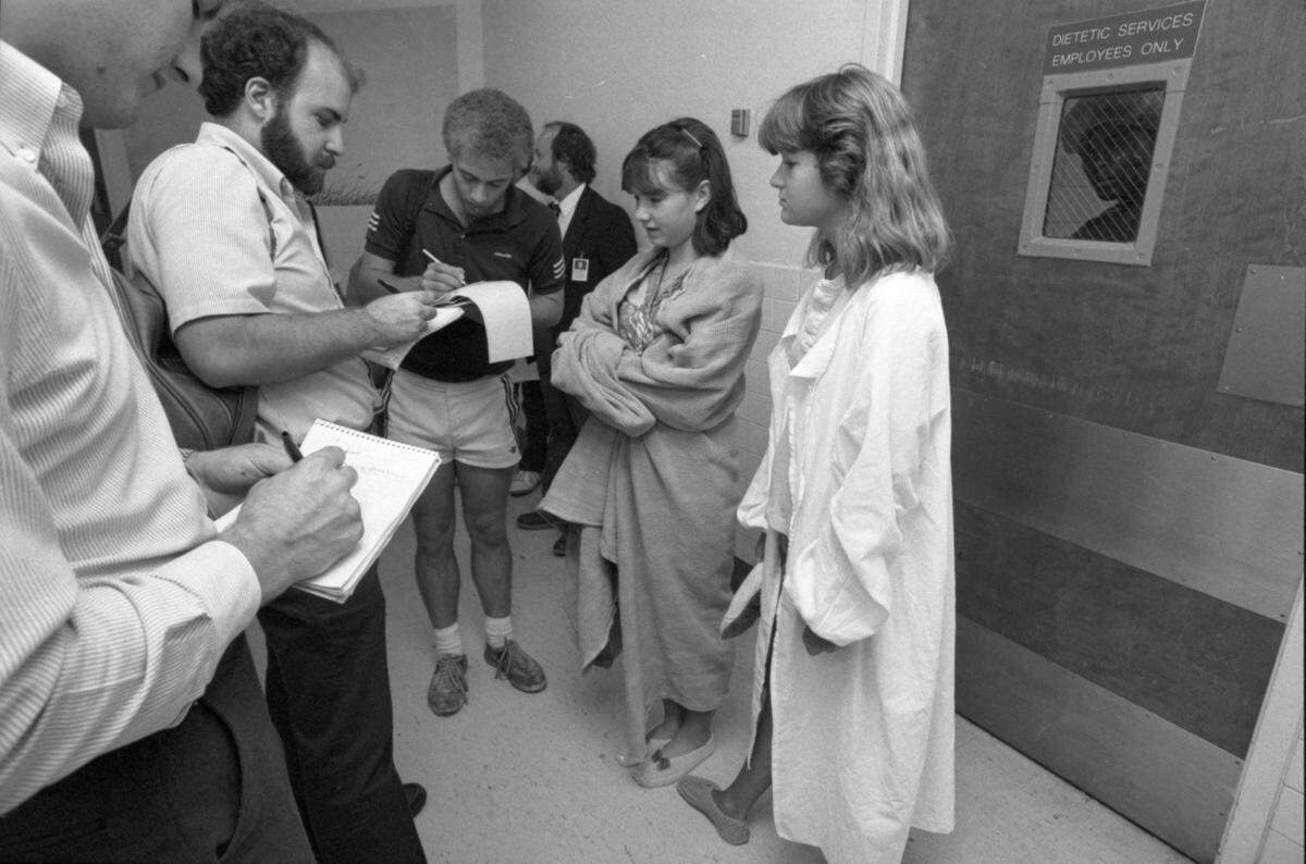 Black and white photo of reporters talking to two young women in the hallway of a hospital.