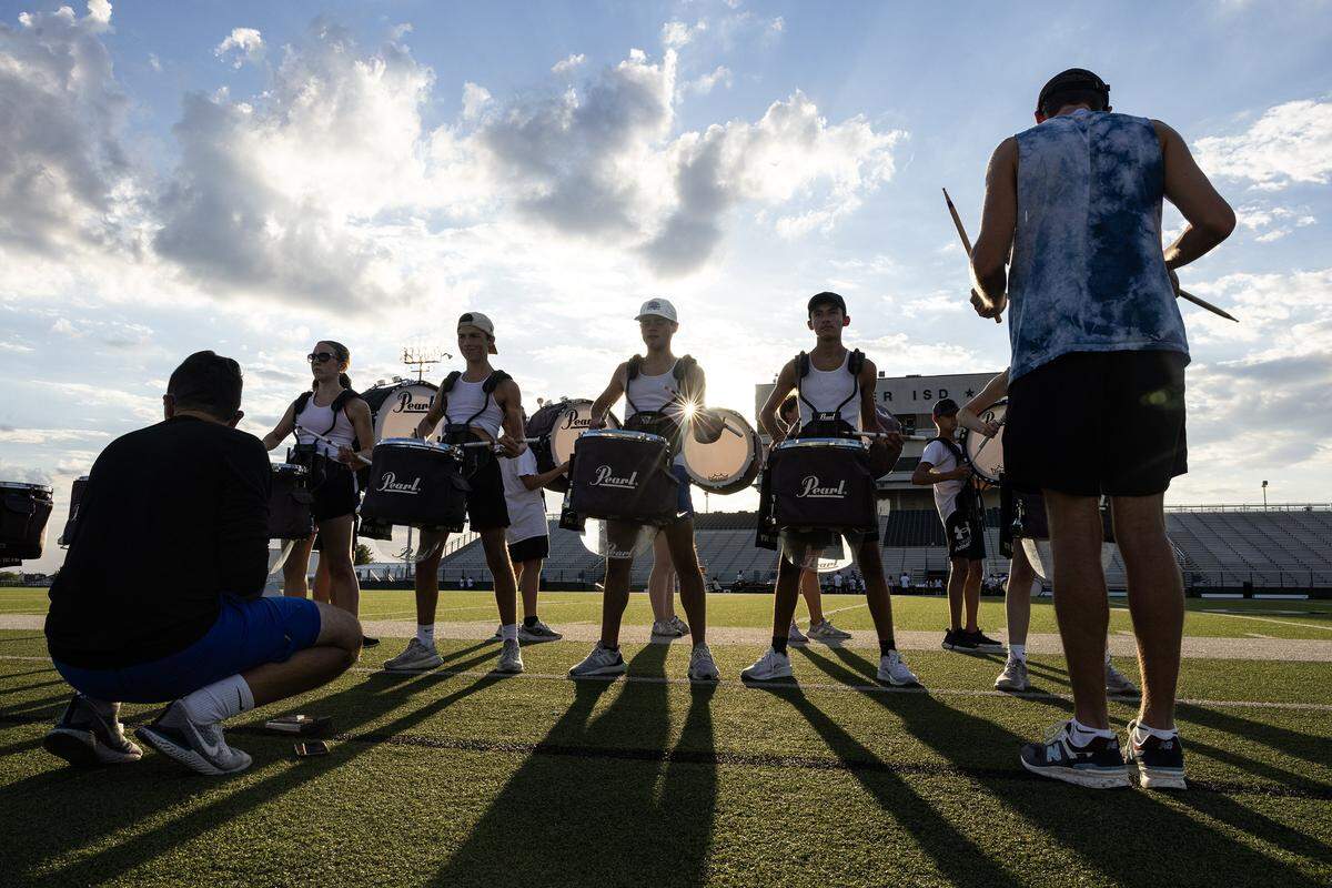 Drum line members with snares and bass drums are backlit as the sun shines behind them, casting a shadow on the football field. 