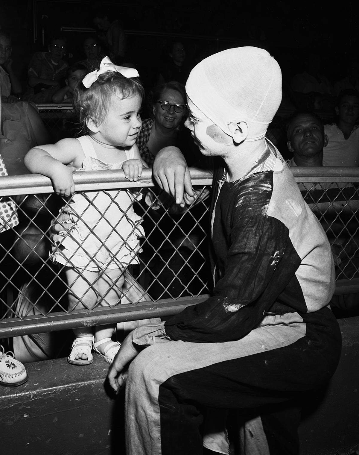 A toddler leaning over a wire fence smiles at a boy dressed like a clown.