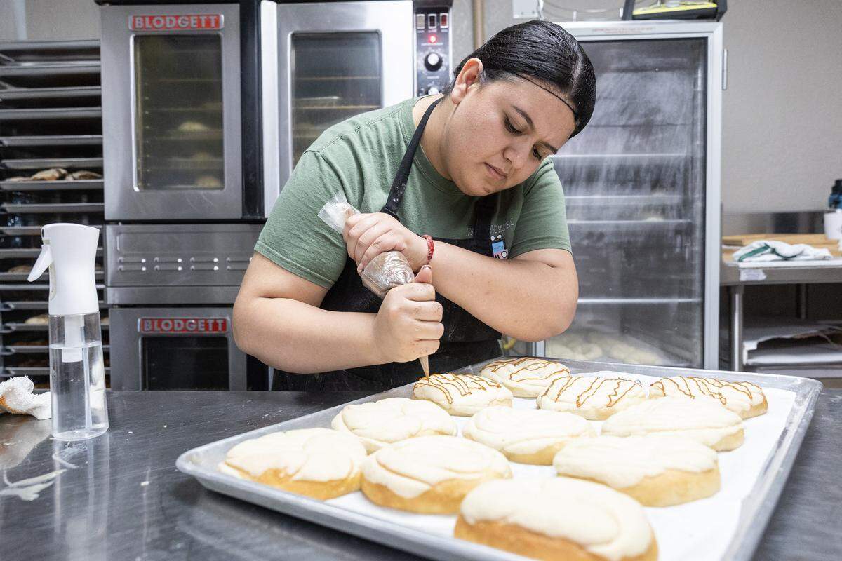 A baker drizzles caramel on top of pastries sitting on a tray in an industrial kitchen.