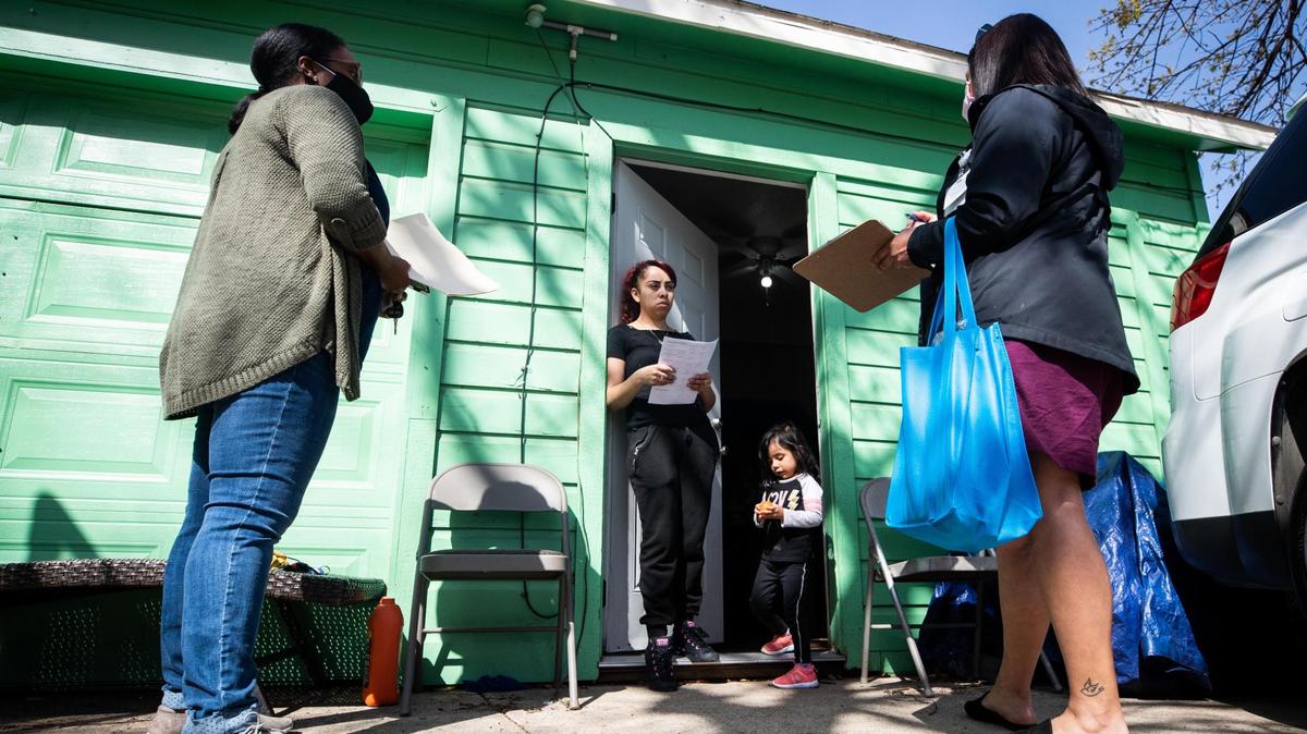 Two masked woman stand in front of a green house and talk to a third woman as a little girl looks on. 