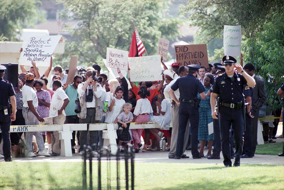 Dallas Mayor Pro Tem Diane Ragsdale, surrounded by Dallas police, protests Queen Elizabeth II’s visit to Dallas.  A large crowd stands behind a barricade holding signs.