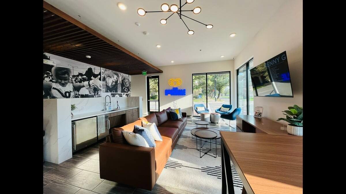 The lobby of an apartment building with a leather couch facing towards a bank of televisions to the right with windows looking out to the street in the background. 