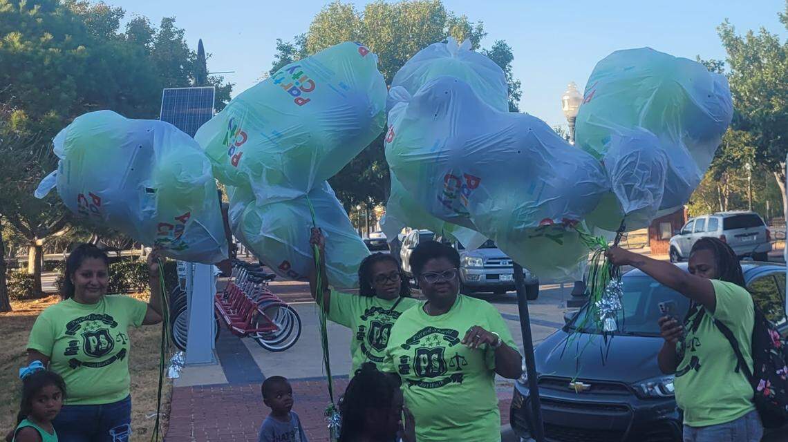 Four women in green Parent Shield T-shirts stand on a sidewalk holding big bags of helium balloons floating over their heads. Three young kids watch curiously.