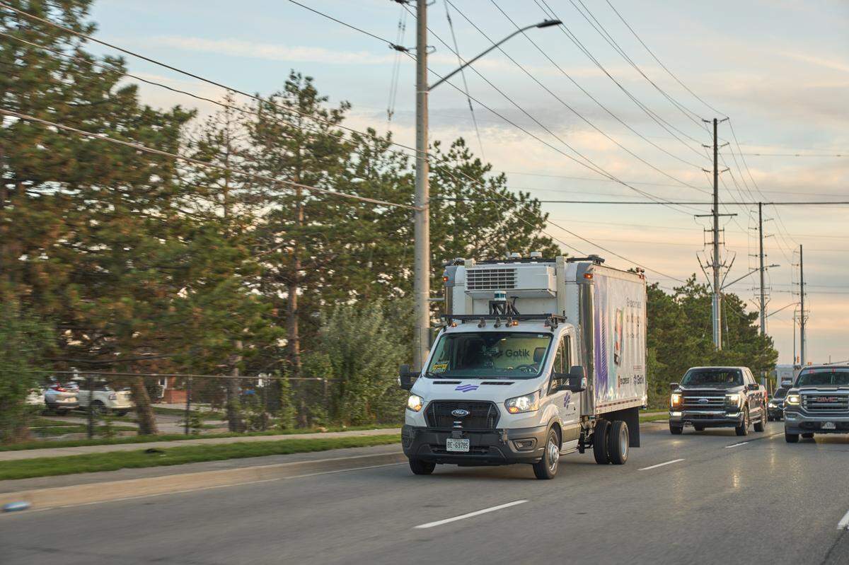 A self-driving truck drives on a road with cars to the right. There is no one in the front seat steering the vehicle.