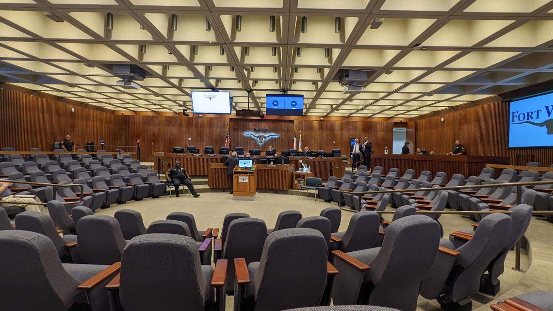 A wide angle of a city council chamber. An 11 member panel is in front surrounded by a semi-circle of four rows of stadium seats.