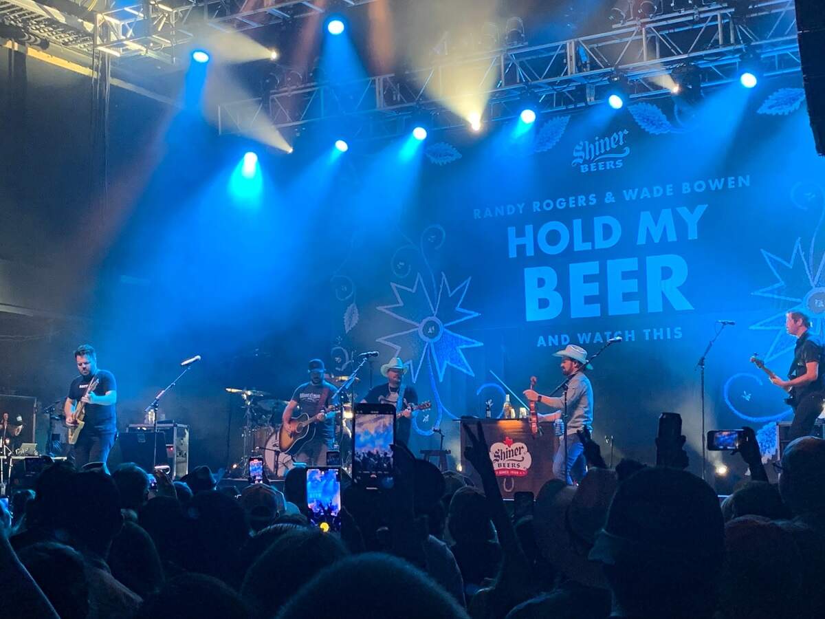 Two male lead singers and a backing band of three other men perform on stage with guitars in front of a backdrop that reads quote Randy Rogers and Wade Bowen: Hold My Beer.