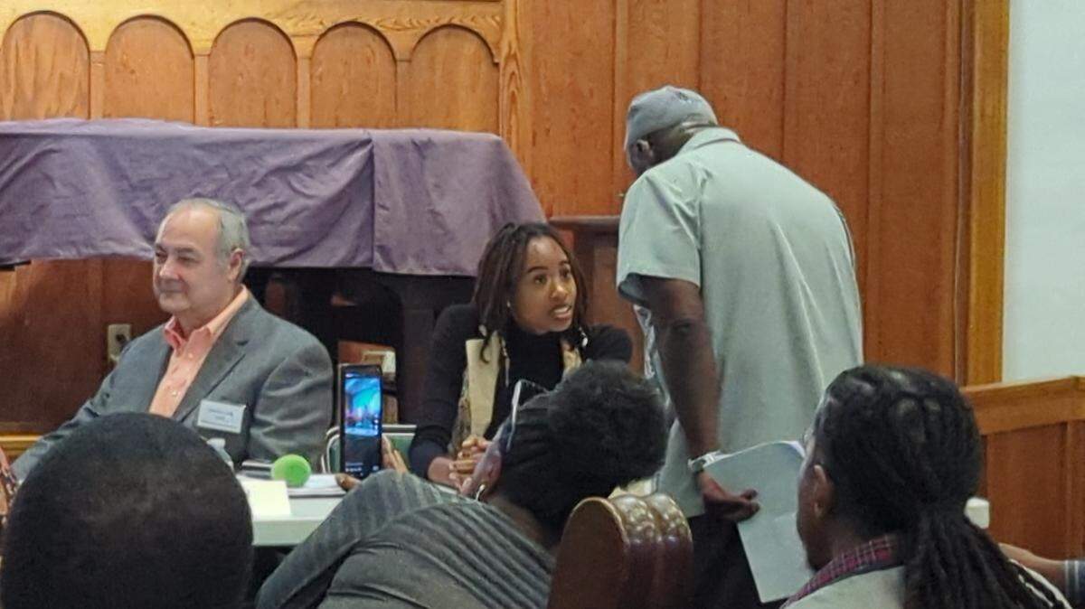 A young black woman with dreds sitting at an event table looking incredulous speaking to an event organizer about why she can't participate in a city council candidate forum. 