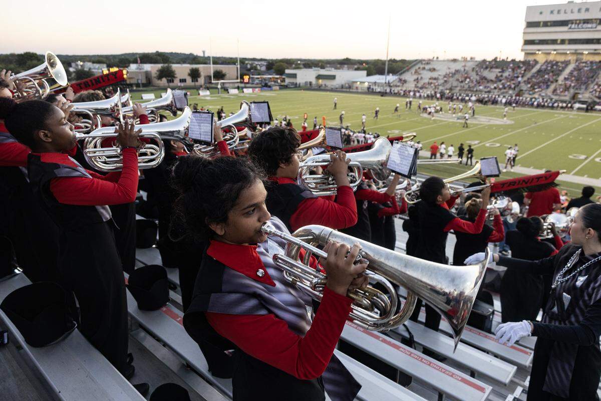 A brass player in the foreground looks to the right while in the stands. Behind her are numerous other band members dressed in black and red uniforms. 