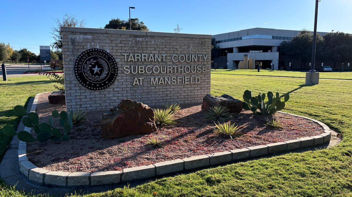 A white brick waist-high sign in a field of green grass features a circular seal and text. Cactuses, succulents and rocks decorate the space in front of the sign. A white building peeks from the background with the blue sky overhead.