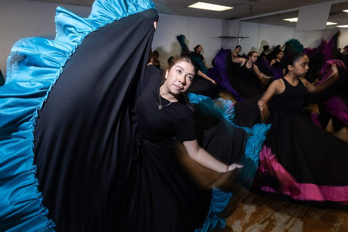 A young woman lifts the skirt of her black skirt with blue trim with her right hand while dancing.
