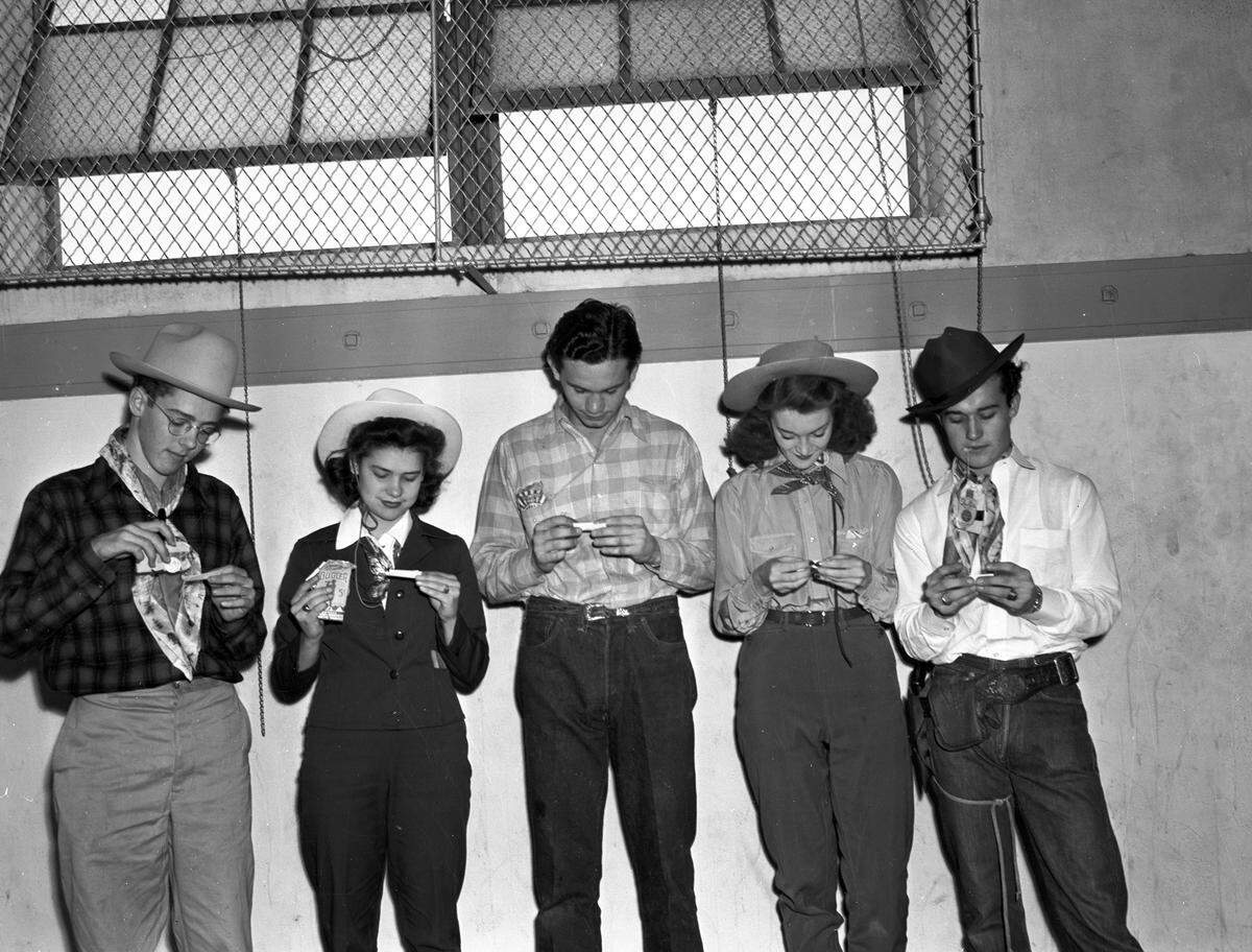 The group of five are standing side-by-side against a wall, each of them wearing stylish Western attire. Bill is rolling a cigarette while he appears to be holding the pack of tobacco with his mouth from a piece of the package. Mr. Radcliff and Elna Jean are both rolling their cigarettes, their tobacco packs appear to be peaking from their pockets. Betty is holding her Bugler pack in one hand and her cigarette in the other, seemingly inspecting it. Edwin is sprinkling some tobacco into his rolling paper.